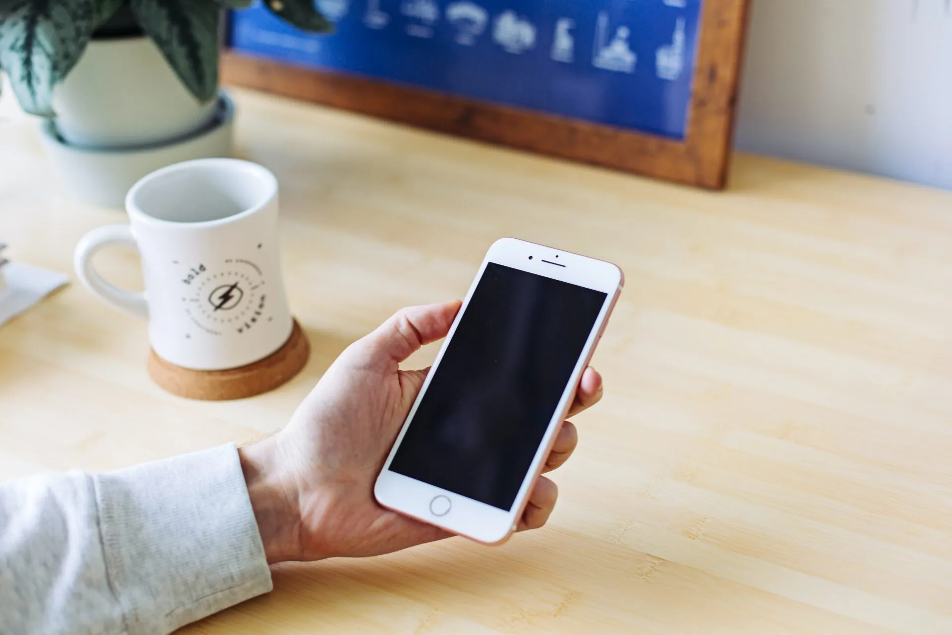 A hand holding a phone beside a coffee mug on a wooden desk